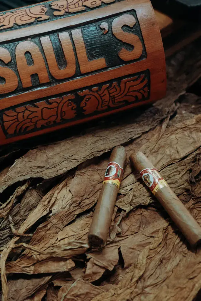 Two cigars resting on tobacco leaves beside a carved Saul's sign