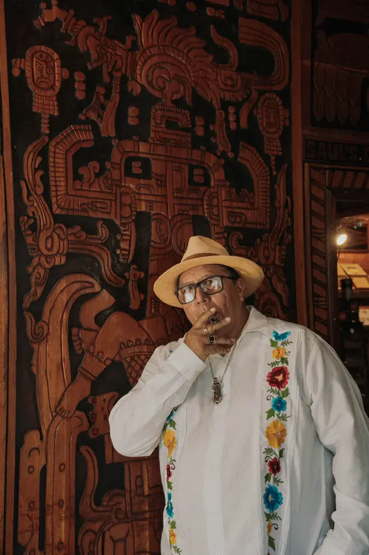 Saul Nuñez in a straw hat and embroidered guayabera, with a cigar, in front of the carved wooden wall of his shop